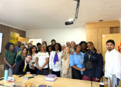 A diverse group of colleagues gathered around a wooden table with a small purple cake and snacks for an office celebration. The group is standing together posing for a photo in what appears to be a meeting or break room with a projector mounted on the ceiling